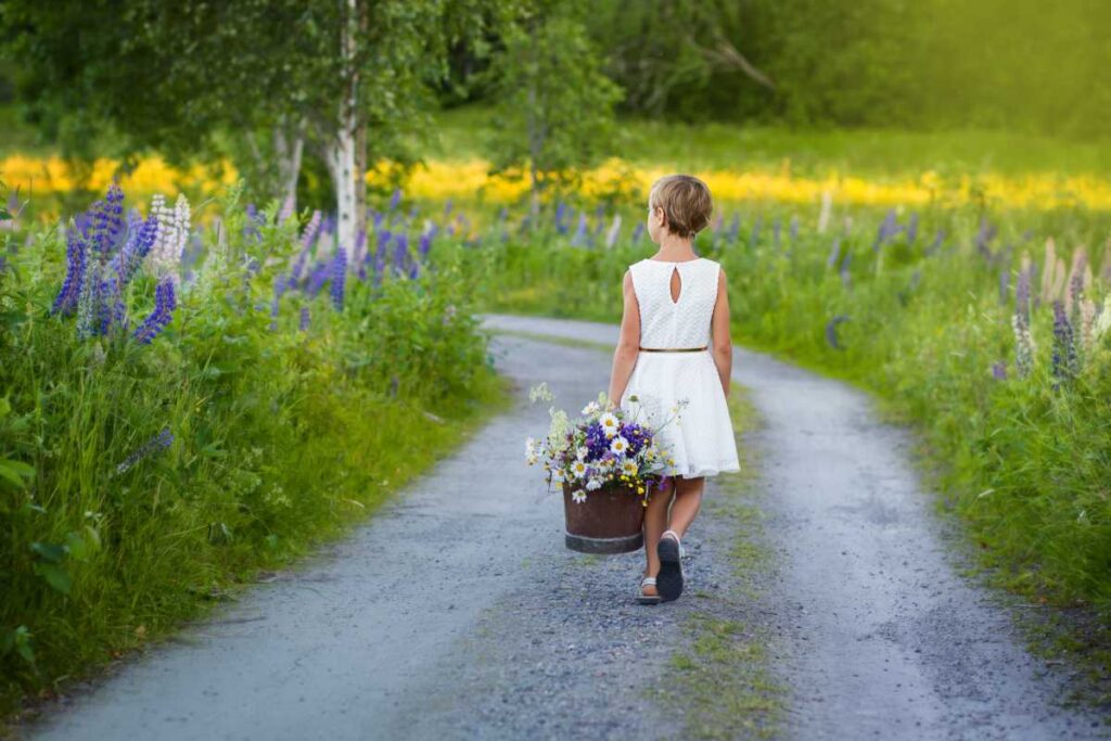 En flicka går på en grusväg med en hink med blommor i handen